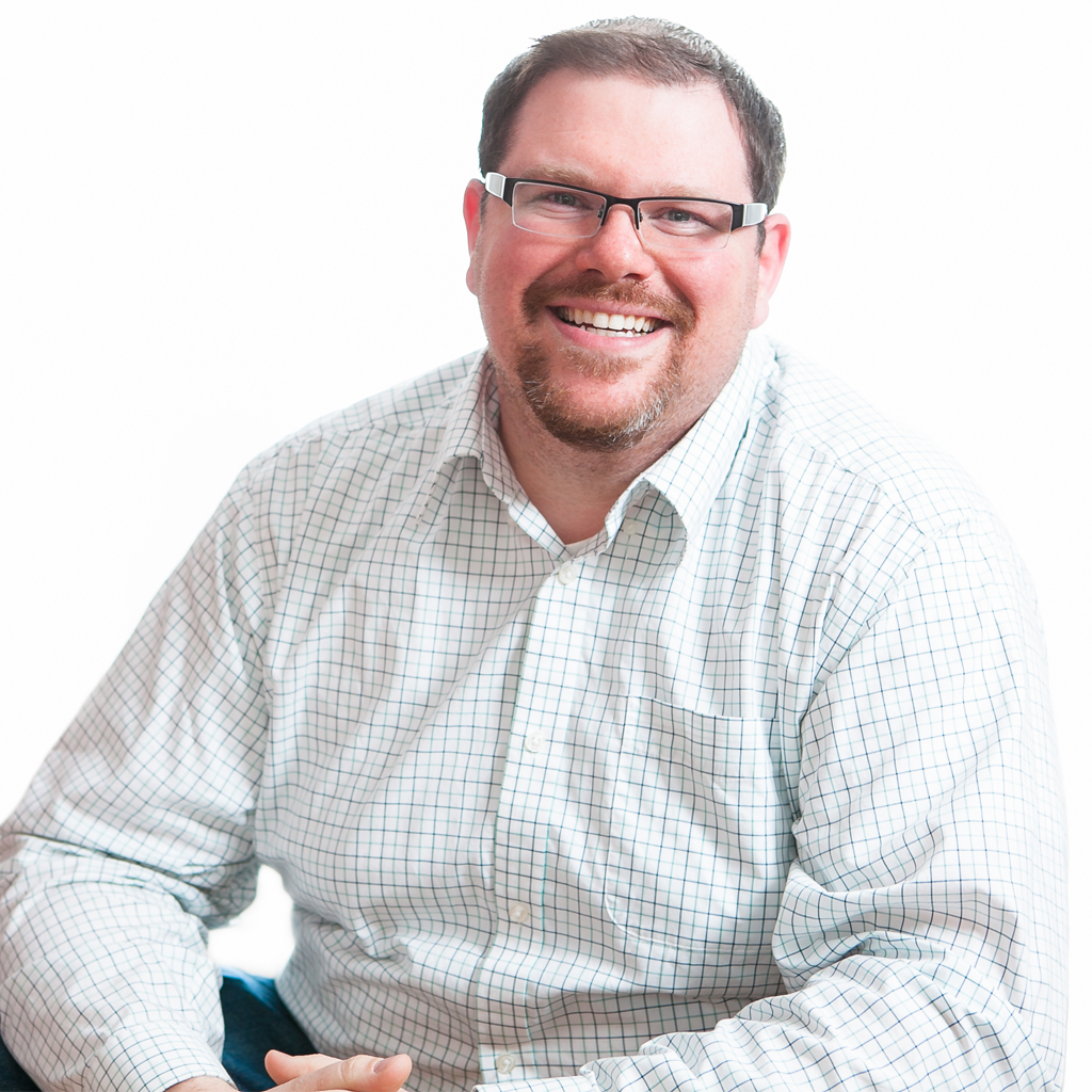 Smiling man (John Picozzi) in a checkered shirt and glasses, seated against a white background.